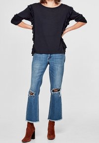 Svart rynkad blus med långa ärmar som matchas med ljusblå slitna jeans. Modellen har på sig rödbruna ankelstövlar med klack.