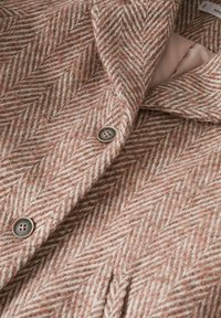 Vlněný kabát v herringbone vzoru, s jemnou kombinací růžové a krémové barvy, kovovými knoflíky a klopovým límcem.