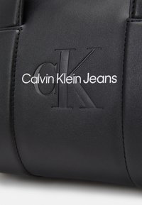 Matière en cuir noir avec un grand logo "cK" embossé et un texte blanc "Calvin Klein Jeans" imprimé dessus, cousu sur le côté.