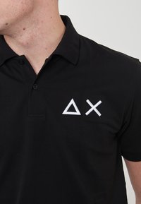 Polo nero con colletto, caratterizzato da forme geometriche ricamate bianche (triangolo e croce) sul lato sinistro del petto. Tessuto texture.