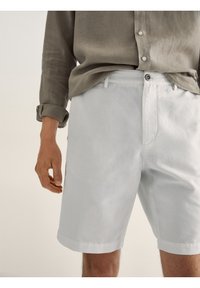 Shorts en lin gris clair avec un devant plat, ourlets roulés et une fermeture à bouton unique. Associés à une chemise olive à coupe ample.