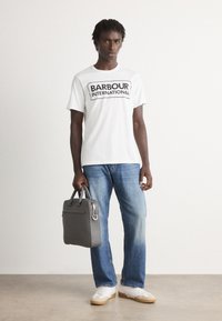 Mladý muž v bílém tričku "Barbour International", modrých džínách a bílých teniskách, který drží šedý kufřík vedle sebe.