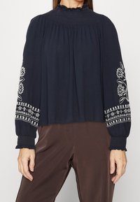 Marineblaue Bluse mit gerafftem Stehkragen, langen Ärmeln mit weißer floralem Stickerei und rüschengesäumten Manschetten, kombiniert mit braunen Hosen.