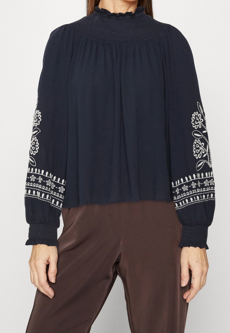 Marineblaue Bluse mit gerafftem Stehkragen, langen Ärmeln mit weißer floralem Stickerei und rüschengesäumten Manschetten, kombiniert mit braunen Hosen.