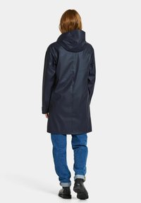 Marinblå vattentät regnrock med huva, slät textur, långa ärmar och rak skärning, kombinerad med blåa denimjeans och svarta skor.