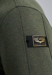 Zelený textúrovaný svetre z látky s čiernou etikettou a zlatým logom, prešité švy a neformálny dizajn.