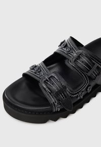 Black sandal with thick sole, two buckle straps, decorative stitching, and embossed floral metal studs on a white background.