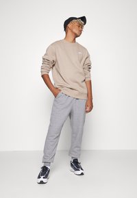 Beige sweatshirt med logotyp, grå sweatpants och svarta sneakers. Modell som bär en svart keps, står i en avslappnad pose med händerna i fickorna.