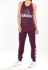 Lilla atletisk tanktop med stort hvidt "adidas" logo, parret med matchende lilla joggerbukser med logo detaljer på siden, iført hvide sneakers.