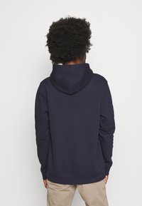 Sudadera con capucha de color azul marino, con una tela texturizada, mangas largas y un bolsillo tipo canguro, exhibida desde la parte posterior sobre un fondo blanco.
