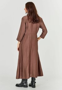 Brown, long-sleeve, ankle-length dress with a relaxed fit. Lightweight textured fabric and subtle gathers at the waist. Paired with black boots.