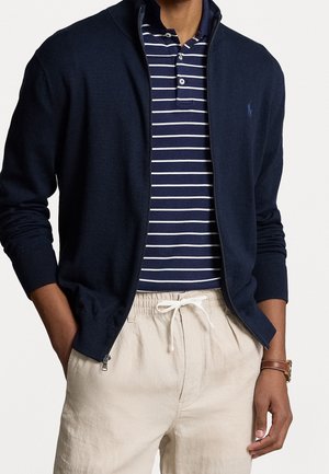 Mężczyzna w granatowej kurtce na zamek, pod którą ma granatowo-białą paskowaną koszulkę polo, beżowe spodnie z troczkami oraz brązowy zegarek na nadgarstku.