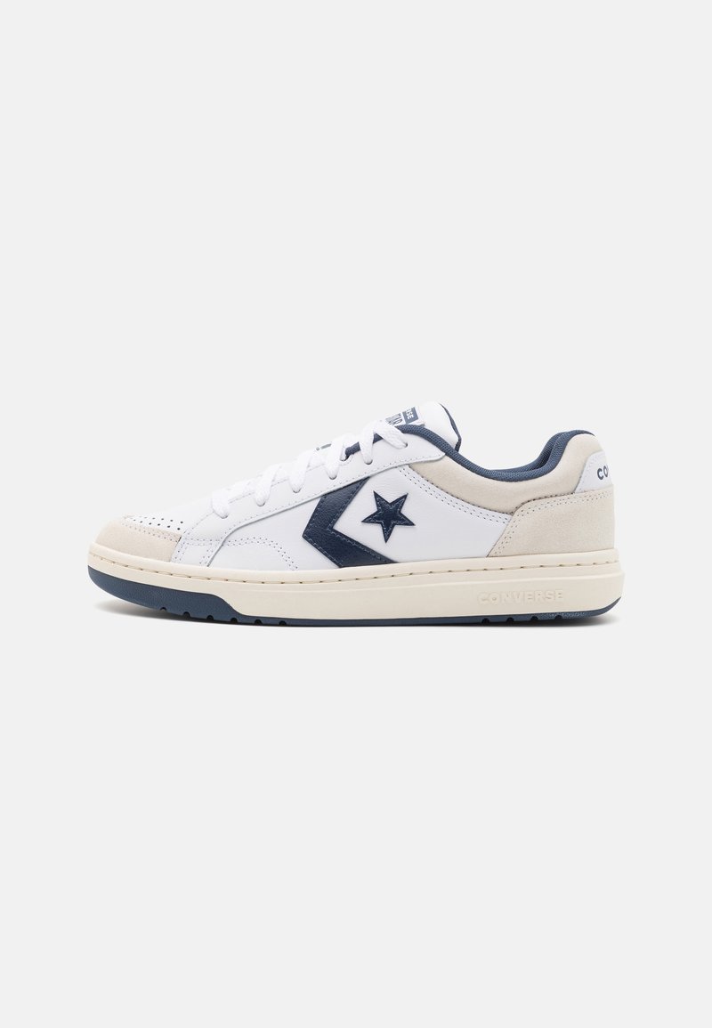 Baskets basses blanches Converse avec logo étoile bleu marine, accents en daim beige sur le bout et le talon, et lacets blancs sur fond gris.