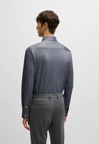 Graues Langarmshirt mit glatter Textur, ausgestattet mit einem Punktkragen und Knopfmanschetten, kombiniert mit grauen maßgeschneiderten Hosen und Gesäßtaschen.