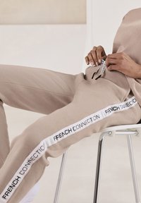 Beige Jogginghosen mit einem weißen Seitenstreifen, auf dem "FRENCH CONNECTION" in Schwarz steht. Hergestellt aus weichem Material, mit elastischem Bund und Kordelzug.