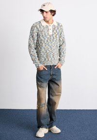Maglione lavorato a maglia blu e crema con colletto e bottoni, abbinato a jeans blu sbiaditi con un ginocchio strappato e sneakers bianche.
