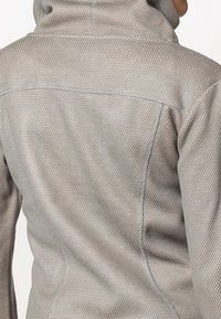 Giacca zip-up grigia chiara con un motivo testurizzato, colletto alto e cuciture a contrasto. Il tessuto sembra liscio e leggermente elasticizzato.