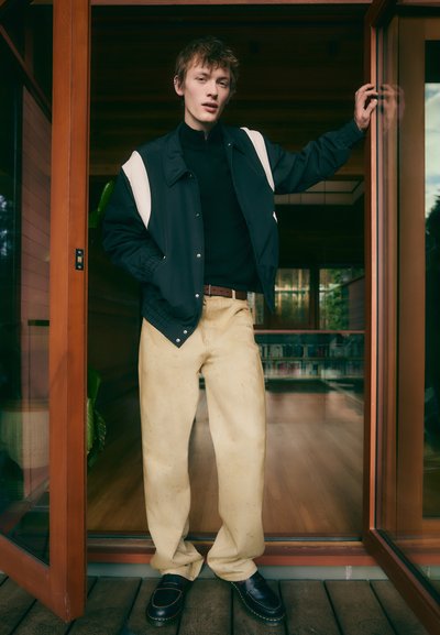 Black zip-up jacket with white accents, black turtleneck, cream-colored trousers, and dark loafers. Natural wood interior visible.