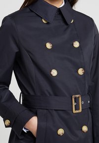 Žena v tmavě modrém dvouřadém trench coat s knoflíky z želvoviny a odpovídajícím opaskem s obdélníkovou sponou, ruka v kapse.