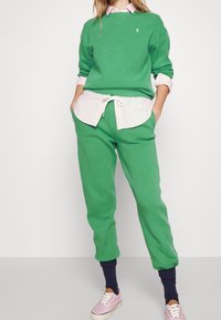 Zelená mikina a odpovídající tepláky vyrobené z bavlny. Přes růžovou košilovou košili. Obsahuje logo na hrudi a žebrované manžety.