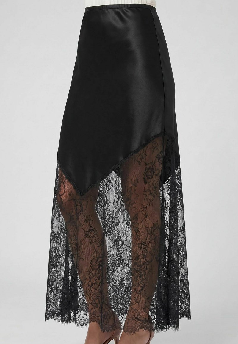 Jupe asymétrique noire avec une couche supérieure unie et un long bas en dentelle florale transparente dévoilant les jambes, portée avec des talons hauts beiges.