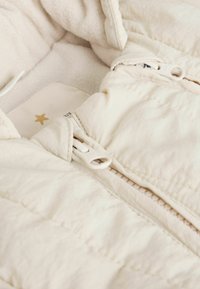 Ljusbeige quiltad jacka med ett mjukt, texturerat innerfoder. Har en hel dragkedja och en liten guldstjärna som detalj på etiketten.