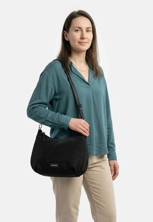 Sieviete ar taisniem brūniem matiem, tērpusies tējas zaļā blūzē un bēšās biksēs, nēsā melnu plecu somu ar redzamu zīmola etiķeti.