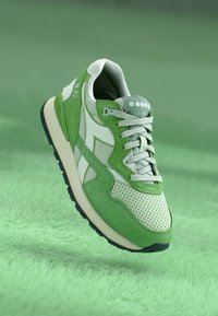 Green sneaker featuring a mix of smooth and perforated materials, with contrasting white accents and a textured grey tongue on a lush background.