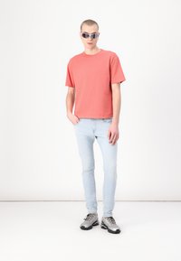 Rosa bomulls T-skjorte, lyseblå jeans, grå joggesko. Modell som har på seg kantete solbriller, står mot en hvit bakgrunn.