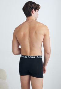 Schwarze Herren-Boxershorts mit einem breiten, weißen, gebrandeten Bund, die eine enge Passform und glattes Material aufweisen, aus der Rückansicht gezeigt.