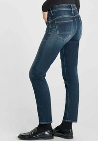 Jean en denim bleu foncé avec une coupe slim, taille haute et design à cinq poches. Présente des coutures claires et des poches arrière avec détail à rabat.