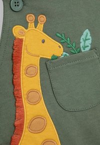 Vêtement en coton vert avec une girafe brodée en jaune, ornée d'accents orange, sur une poche décorée de détails en feuilles vertes.