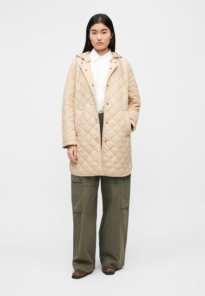 Μπεζ καπιτονέ μπουφάν με κουκούλα, κουμπιά με τσικ, και πλαϊνές τσέπες, φορεμένο πάνω από λευκό πουκάμισο και ελιάς πράσινο παντελόνι cargo. Καφέ παπούτσια.