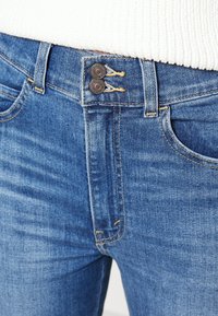 Vaqueros de denim en azul medio, con una cintura alta, cierre de dos botones y detalles de costura amarilla en los bolsillos. Textura suave y acabado liso.