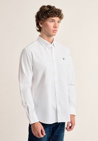 Camisa blanca de manga larga con cuello abotonado, que presenta un pequeño logo bordado en el pecho y un sencillo cierre de botones.