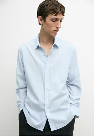 Giovane uomo con capelli corti castani che indossa una camicia azzurro chiaro con bottoni, con le mani nelle tasche dei pantaloni neri, guarda verso il basso.