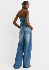 Salopetă din denim cu top fără bretele și pantaloni evazați, având o textură decolorată și studs decorative pe laturi.