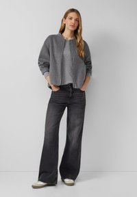 Woman with long blonde hair wearing a gray zippered jacket, gray shirt, wide-leg black jeans, and beige sneakers standing with hands in pockets.