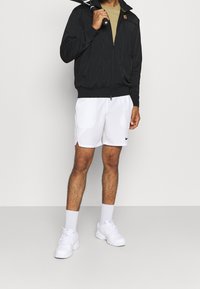 Man i svart jacka, beige skjorta, vita Nike shorts, vita strumpor och vita sneakers som håller en tennisk racket över axeln.