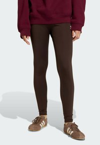 Smeđe tajice od rastezljivog materijala, s glatkom teksturom i prilagođenim dizajnom, uparene s tamnosmešnim sportskim cipelama.