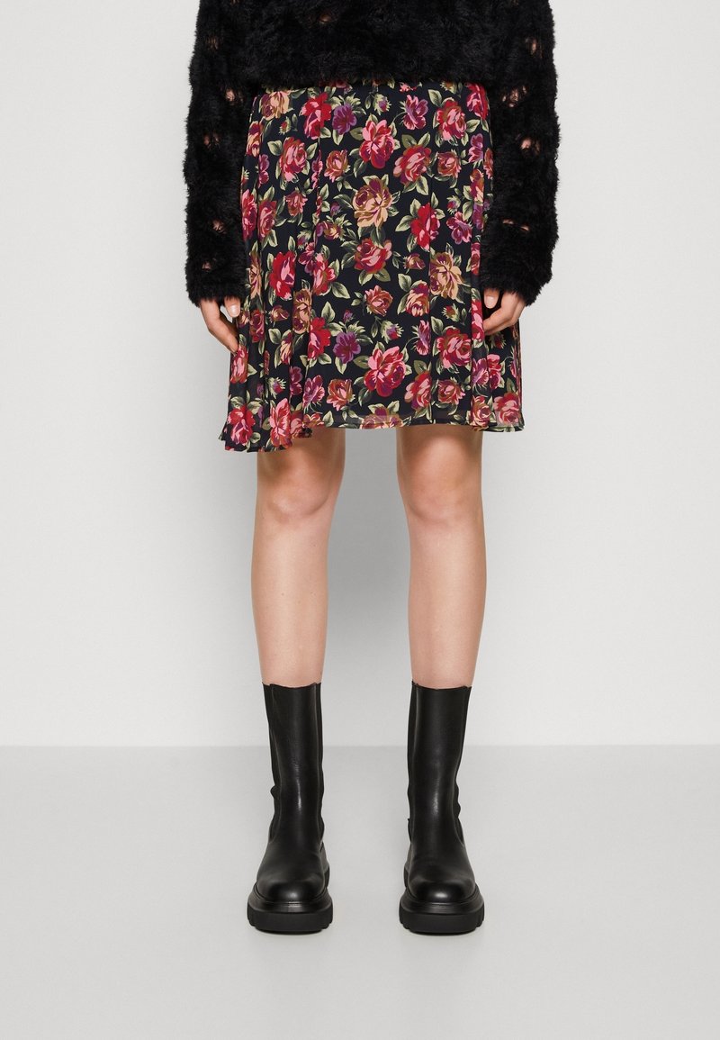 Femme portant un pull noir duveteux, une jupe fleurie jusqu’aux genoux avec des roses rouges et roses, et des bottes en cuir noires mi-mollet, debout sur un sol gris.
