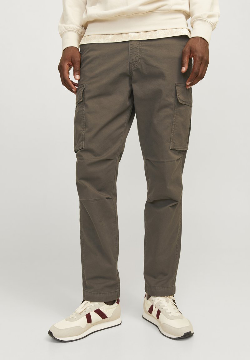 Cargo broek in olijfgroen katoen met zijzakken, taps toelopende pijpen en versterkte stiksels. Gecombineerd met beige sneakers met rode accenten.