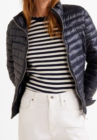 Doudoune bleue marine avec matelassage vertical, fermeture éclair et col haut portée sur un t-shirt à manches longues noir et blanc rayé, associée à un jean blanc.