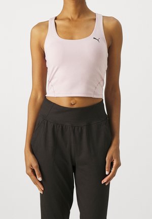 Vrouw draagt een lichtroze cropped tanktop met Puma-logo en zwarte, hooggetailleerde sportbroek tegen een effen achtergrond.