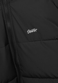 Giacca a vento nera con superficie texturizzata, chiusura con zip e logo argentato. Il tessuto appare liscio e isolato.