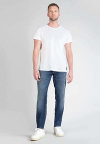 Camisa blanca de manga corta, jeans azul claro de corte ajustado con desgaste, y zapatillas blancas. El modelo se encuentra erguido frente a un fondo neutro.