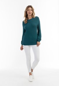 Maglione a maglia testurizzata di colore teal con maniche lunghe e arricciate e scollatura rotonda, abbinato a pantaloni bianchi slim-fit e sneakers bianche.