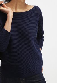 Námořnický modrý pletený svetr s širokým výstřihem, s vodorovným žebrováním a tříčtvrtečními rukávy, nošený s tmavými džíny.