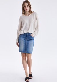 Helles beige Langarmshirt mit strukturiertem Muster, kombiniert mit einem dunkelblauen Jeansbleistiftrock, der über eine frontale Tasche und einen dezenten Saum verfügt.