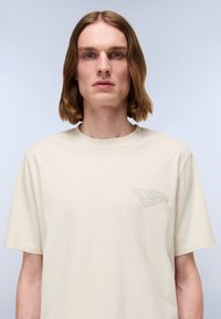 Beige kortærmet T-shirt lavet af bomuld, med et diskret prægget logo på venstre bryst og rund halsudskæring. Glat tekstur.